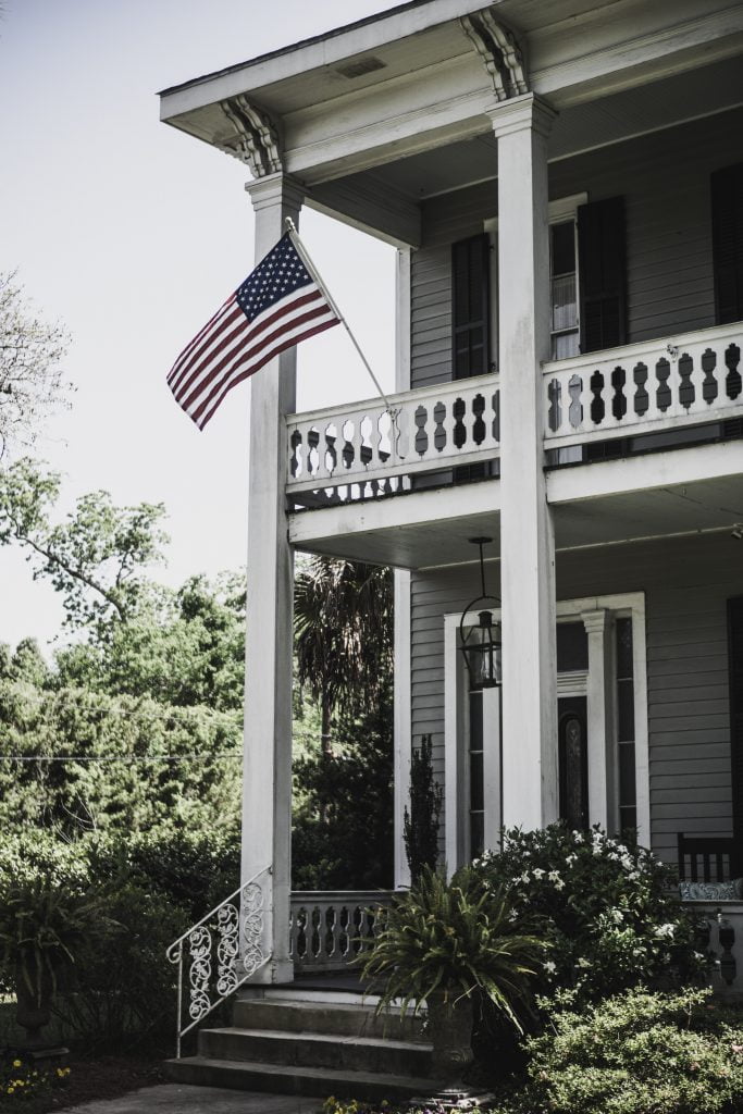 The front porch of a house with the American flag on the second floor balcony.