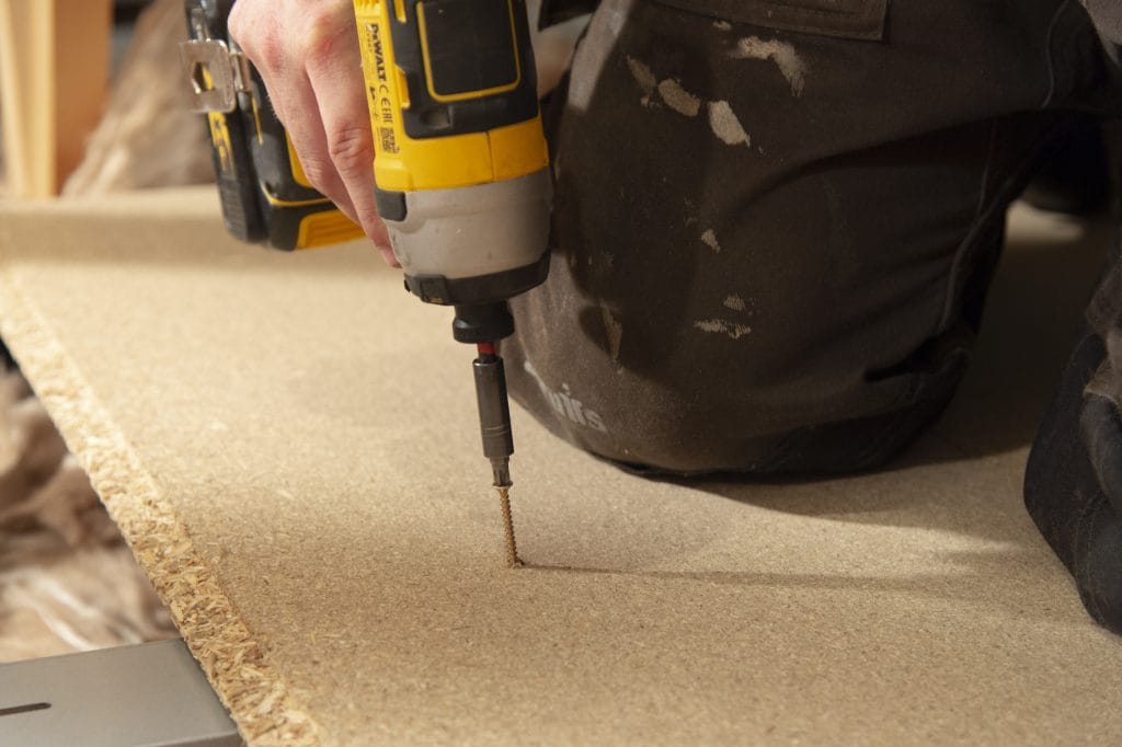 A technician drilling floorboards down in an attic with an electric drill.