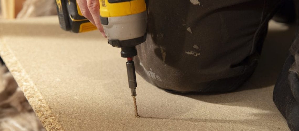 A technician drilling floorboards down in an attic with an electric drill.